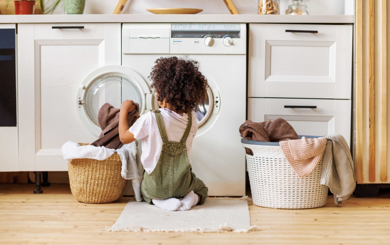 Achteraanzicht van een zwarte jongen met krullend haar die op de grond knielt en kleren in een wasmachine stopt.